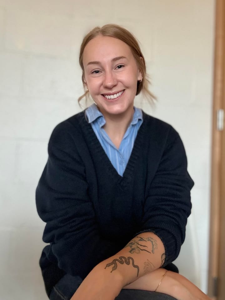 Bailey Pearson, smiling, seated with arms crossed on one knee, wearing a navy sweater over a blue striped shirt